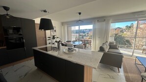 Una cocina moderna con una isla central, un área de comedor con mesa y sillas, y un sofá junto a un ventanal con vistas a la ciudad.