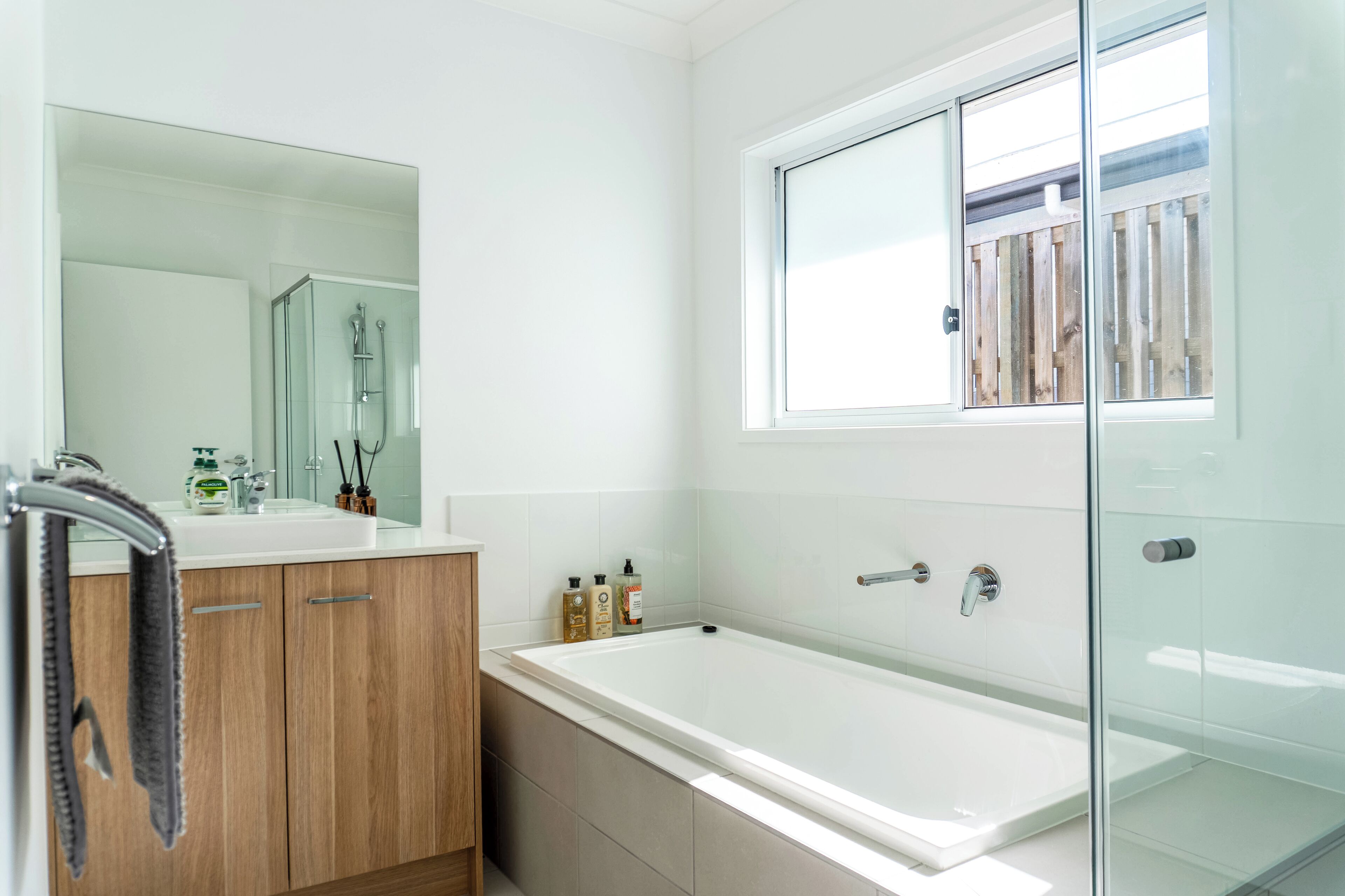 A modern bathroom with a glass shower enclosure, a large mirror, and a window with a view of a wooden fence.