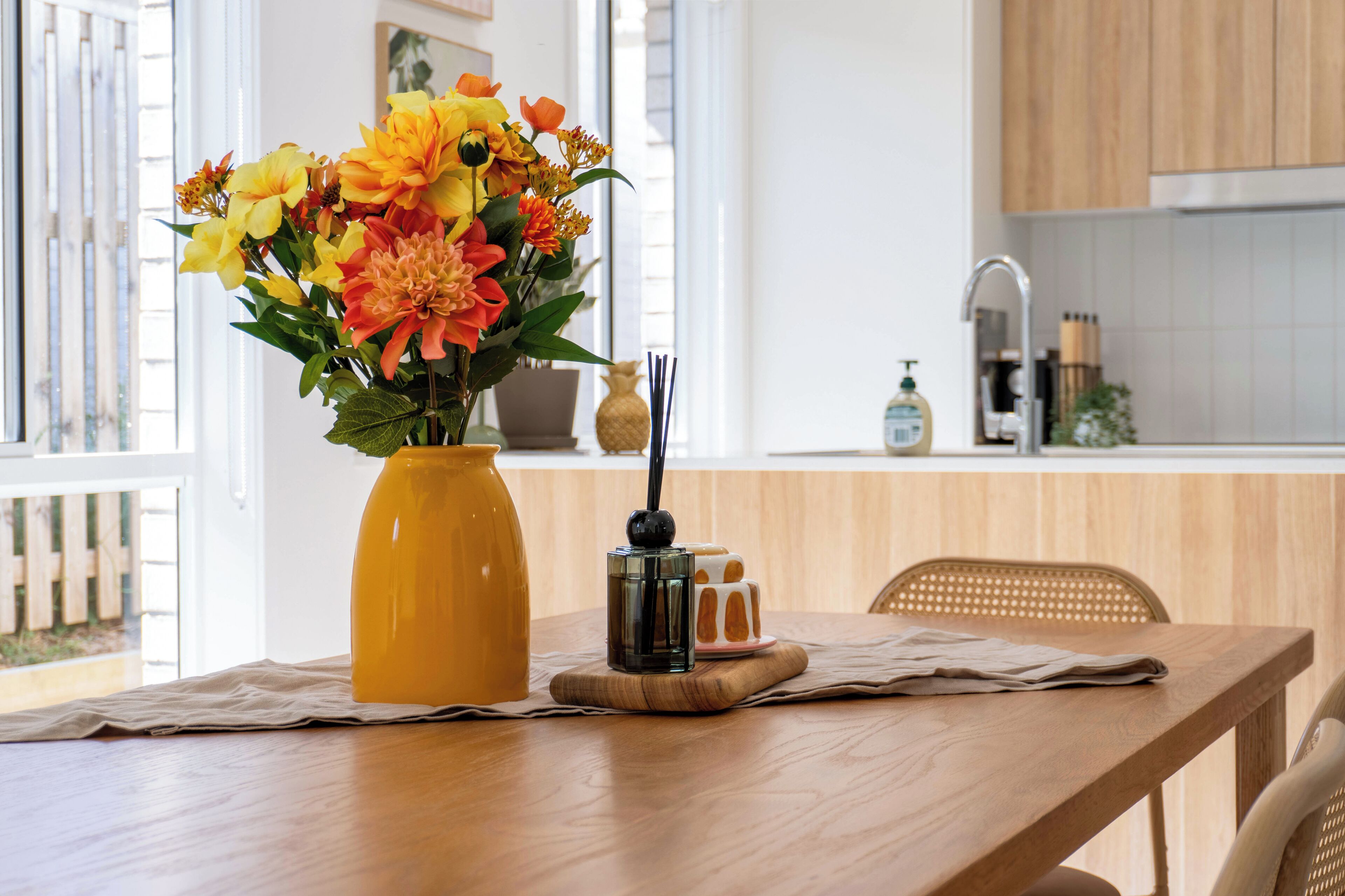 A wooden table with a yellow vase of flowers, a small black container, and a glass of water.
