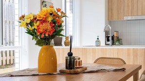 Une table en bois sur laquelle se trouvent un vase jaune rempli de fleurs, un petit récipient noir et un verre d’eau.