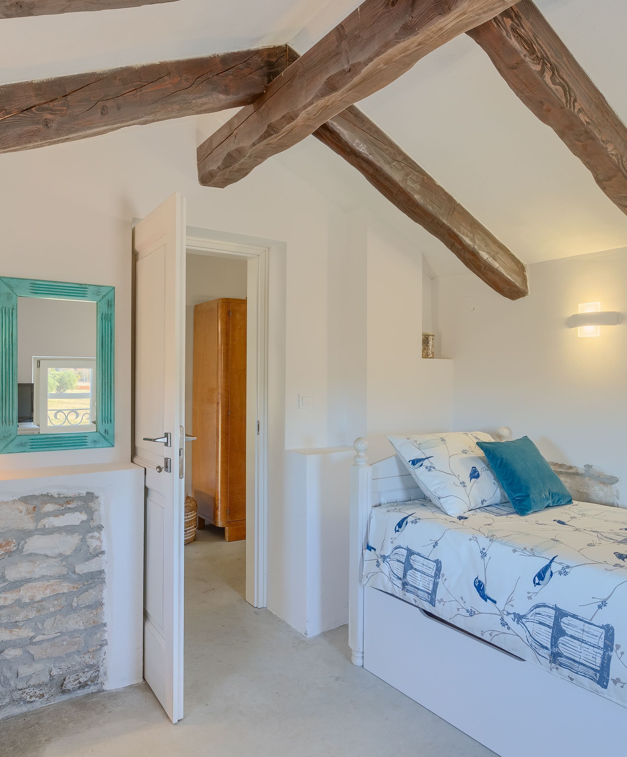 A bedroom with a bed, a mirror, a door leading to another room, and a stone wall.