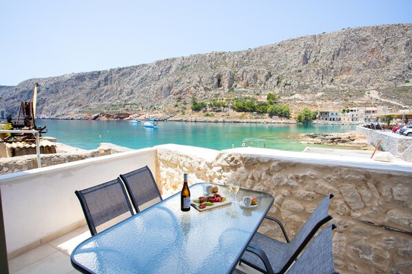 A glass table with chairs, a bottle, and a plate of food on a balcony overlooking a body of water and rocky hills. - Gerolimin Captain Antonis (East Mani)
