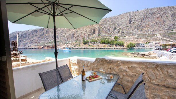 A glass table with chairs and an umbrella, overlooking a coastal area with a boat and buildings. - Gerolimin Captain Antonis (East Mani)