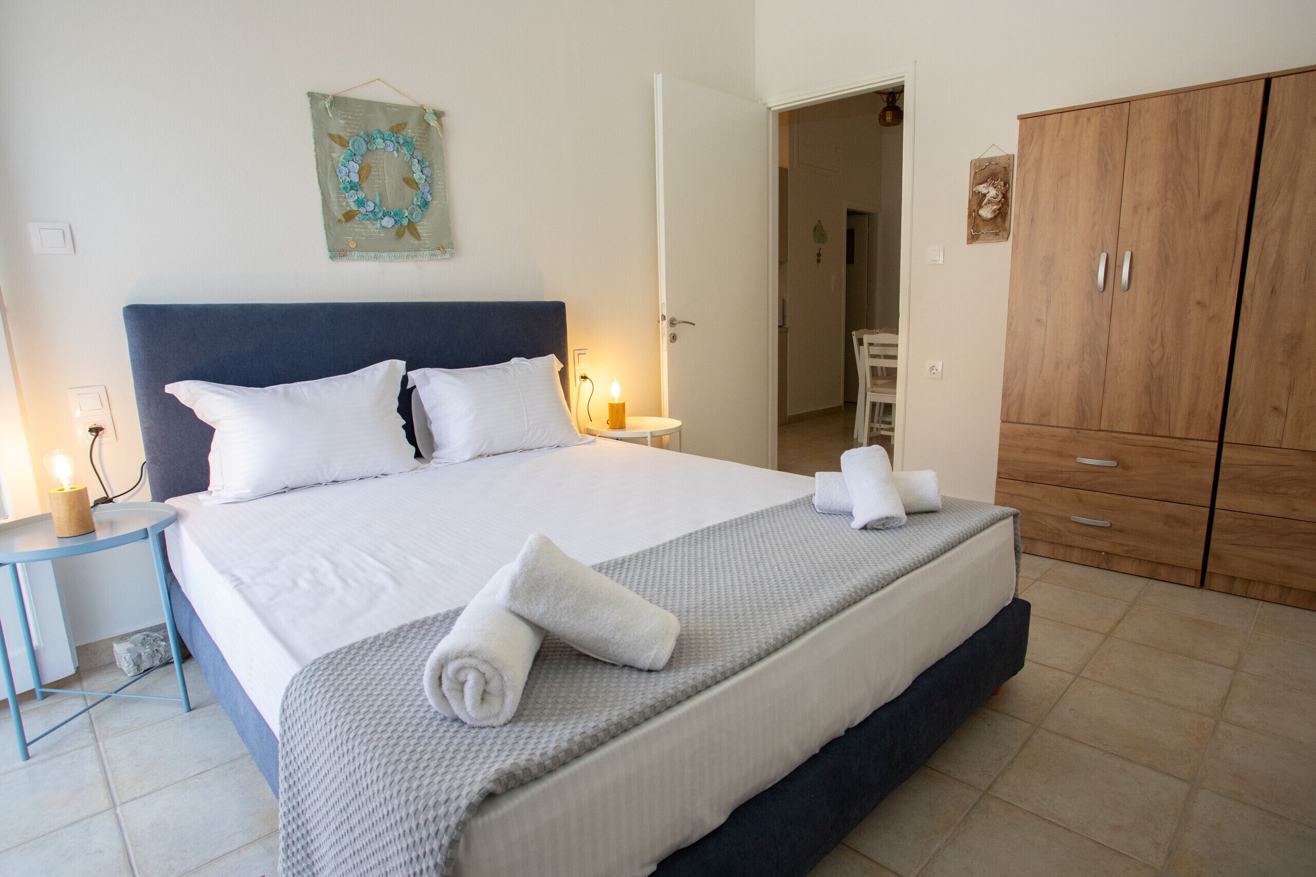 A neatly made bed with white linens and a grey blanket, a wooden wardrobe, and a small bedside table with a candle.