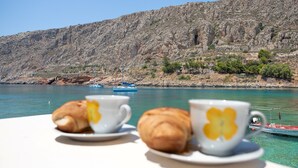 A table with croissants and coffee cups, overlooking a body of water and a rocky hillside. - Gerolimin Captain Antonis (East Mani)