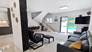 A modern living room with a black sofa, a coffee table, and a dining area with chairs. There is a staircase leading to a balcony with a pool view. - Two-storey house with pool in village of Štrika (Umag)