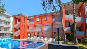 A multi-story hotel with orange and white exterior walls, a swimming pool, and sun loungers. - Bright home with covered porch (Bibione)