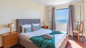 Een netjes ingerichte slaapkamer met een bed, nachtkastjes, een stoel en uitzicht op zee door gordijnen.