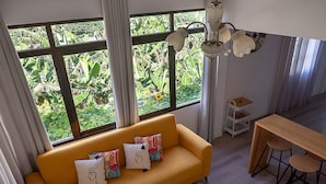 Ein Zimmer mit einem gelben Sofa, einem Esstisch mit Hocker und Blick auf Grün hinter großen Fenstern.