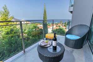 A balcony with a wicker table, a bottle of wine, and a glass, overlooking a coastal town. - LAS ROSAS vista Baia do Funchal (Funchal)