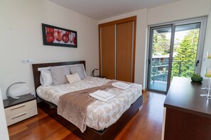 A modern bedroom with a large bed, a bedside table, a nightstand, a painting, a sliding door to a balcony, and a view of greenery outside. - LAS ROSAS vista Baia do Funchal (Funchal)