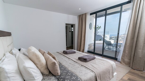 Quarto moderno com uma cama grande, vista da cidade através de uma porta de correr de vidro e uma varanda com mobiliário exterior.
