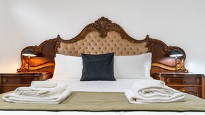 Una cama bien tendida con cabecera capitonada, dos lámparas de noche y toallas dobladas sobre una manta beige.