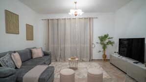 Een woonkamer met een grijze bank, poufs, een klein rond tafeltje, een televisie op een kast en een groot raam met gordijnen.