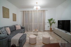 Ein Wohnzimmer mit grauem Sofa, Poufs, einem kleinen runden Tisch, einem Fernseher auf einer Anrichte und einem großen Fenster mit Vorhängen.