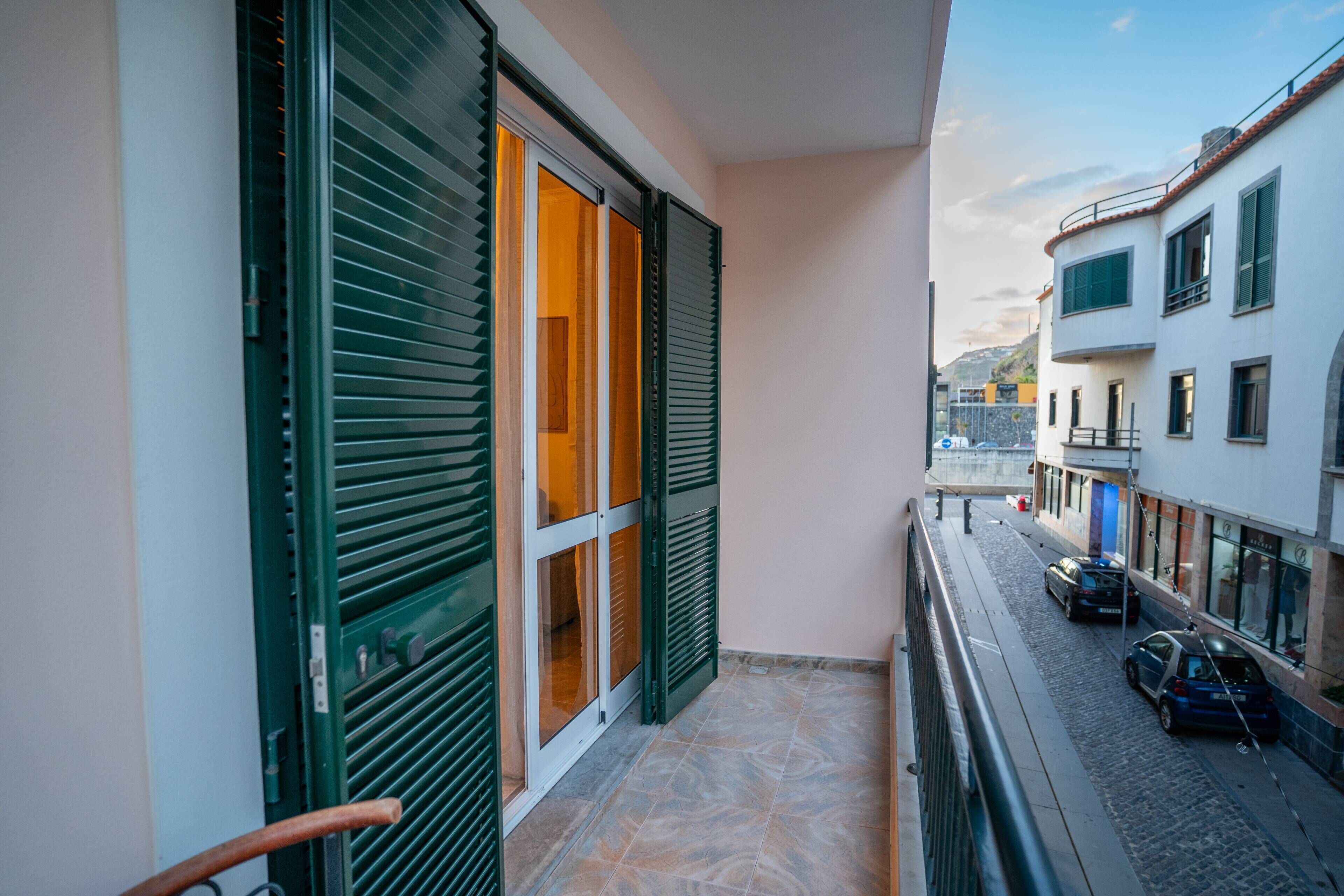A narrow balcony with green shutters, a tiled floor, and a view of a street with parked cars and buildings.