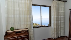 A bedroom with a wooden dresser, a bed with a patterned bedspread, and a window with a view of a coastal landscape.