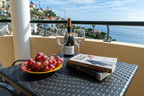 Ein Tisch mit einer Weinflasche, zwei Gläsern, einer Schüssel mit Trauben und zwei Büchern auf einem Balkon mit Blick auf die Küste.