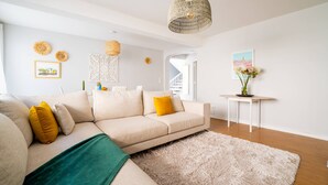 Een moderne woonkamer met een beige bank, een groene plaid en een wit vloerkleed. Er staat een klein wit tafeltje met een bloemenvaas.