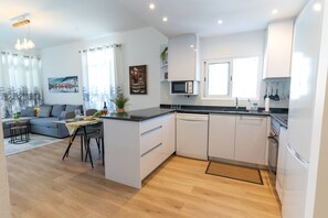 Moderni keittiö, jossa on valkoiset kaapit, sisäänrakennettu mikroaaltouuni sekä ruokailutila, jossa on harmaa sohva ja pieni pöytä.
