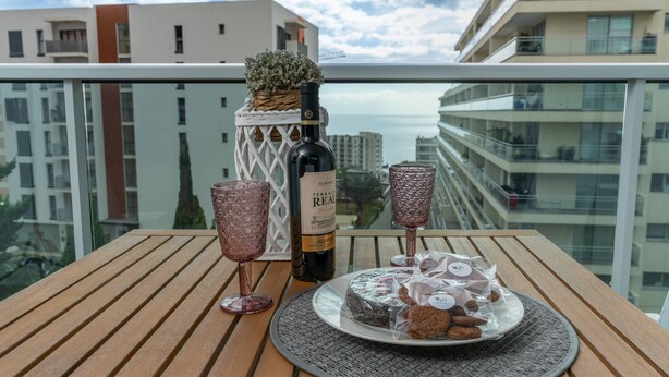 Een balkon met een houten tafel, een fles wijn, twee glaasjes, en een bord met cake en koekjes.