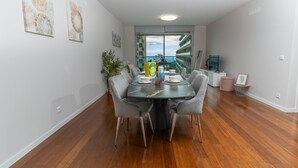 Een moderne eetruimte met een grote tafel, grijze stoelen en uitzicht op de zee door een glazen deur.