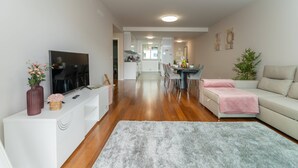 Een moderne woonkamer met een flatscreen televisie, een witte entertainmentkast, een grijs vloerkleed, een beige bank en een eethoek met tafel en stoelen.