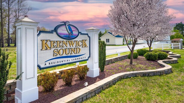A residential community sign with a bird logo, surrounded by well-maintained landscaping and a tree. - Located in Keenwick Sound with Backyard Oasis: fire pit, bar, & TV; Community Pool & Boat/Kayak launch (Selbyville)