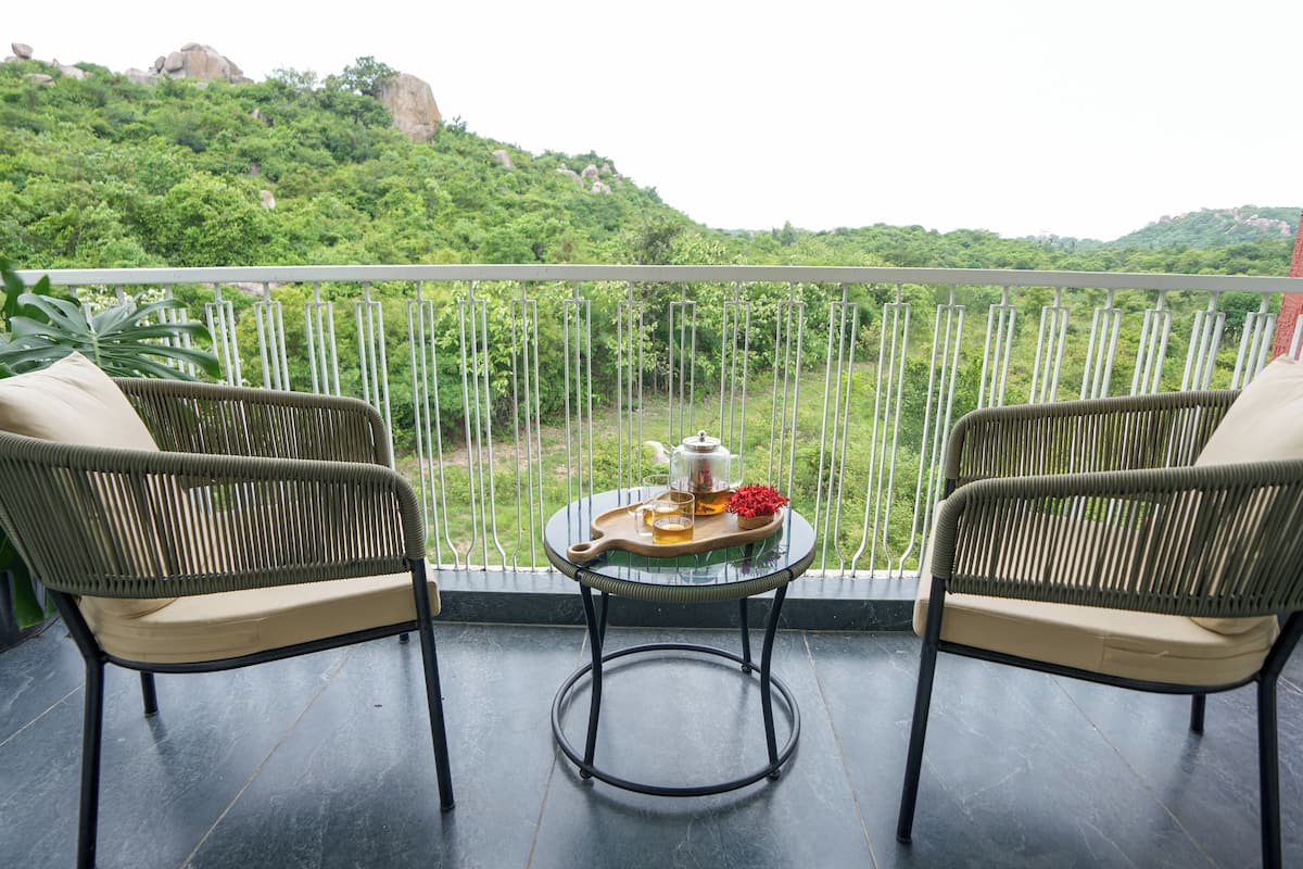 A balcony with two wicker chairs, a small table with a teapot, glasses, and a tray of tea, overlooking a lush green landscape.