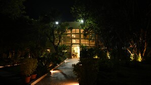 Une vue nocturne d’un bâtiment dont l’entrée est bien éclairée, entouré de verdure.