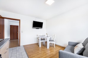 A modern living room with a flat-screen TV mounted on the wall, a dining area with a table and chairs, and a grey sofa with a decorative pillow. - Apartment 'Small Holiday  3 Monteurs' with Shared Garden and Wi-Fi (Weddingstedt)