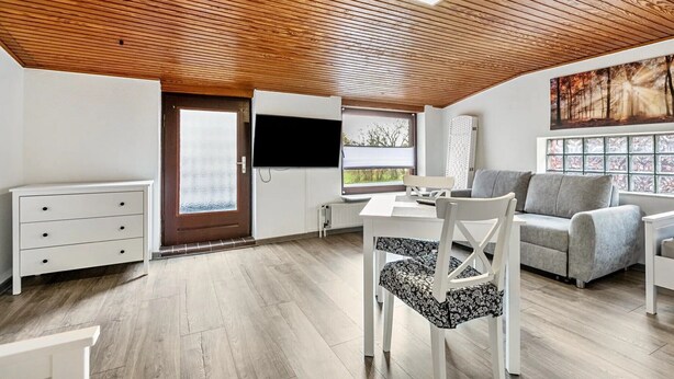 A modern interior with a wooden ceiling, a dining area with a white table and chairs, a grey sofa, and a large window with a view. - Apartment 'Small Holiday  3 Monteurs' with Shared Garden and Wi-Fi (Weddingstedt)