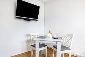 Ein weißer Esstisch mit zwei Stühlen, ein an der Wand befestigter Flachbildfernseher und ein kleiner Tisch mit einer Flasche und einer Fernbedienung.