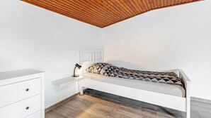 A single bed with a geometric-patterned pillow, a bedside lamp, a white dresser, and a wooden ceiling. - Apartment 'Small Holiday  3 Monteurs' with Shared Garden and Wi-Fi (Weddingstedt)
