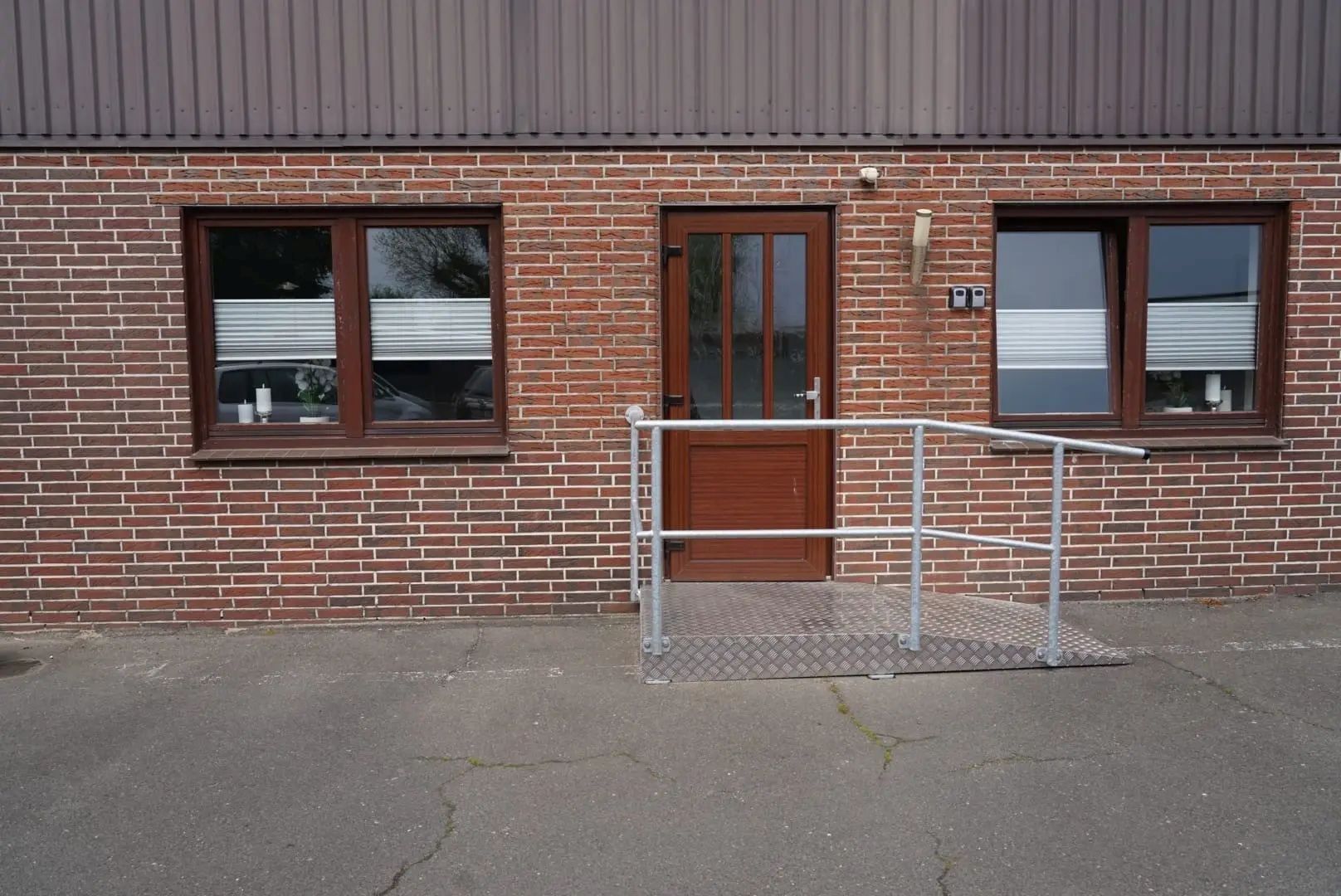 Un bâtiment en briques avec une rampe métallique menant à une porte, une fenêtre avec des stores et un garde-corps en métal.
