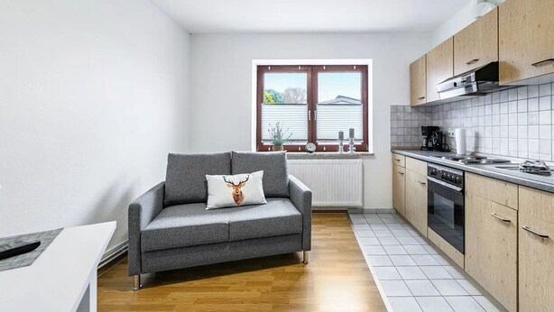 A compact kitchen with a grey sofa, wooden cabinets, and a window with blinds. - Apartment "Practical Mechanic Room 5" with Shared Garden and Wi-Fi (Weddingstedt)