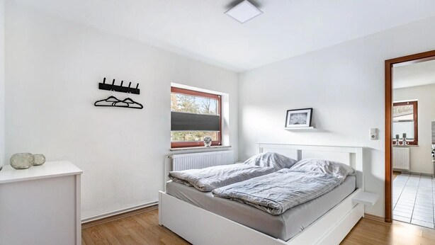 A bedroom with a white bed, a small bedside table, a window with blinds, a radiator, and a wall-mounted coat rack. - Apartment "Practical Mechanic Room 5" with Shared Garden and Wi-Fi (Weddingstedt)