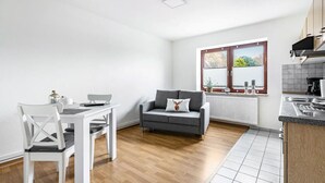 A modern kitchen with a stove, a dining table with chairs, a grey sofa, and a window with blinds. - Apartment "Practical Mechanic Room 5" with Shared Garden and Wi-Fi (Weddingstedt)