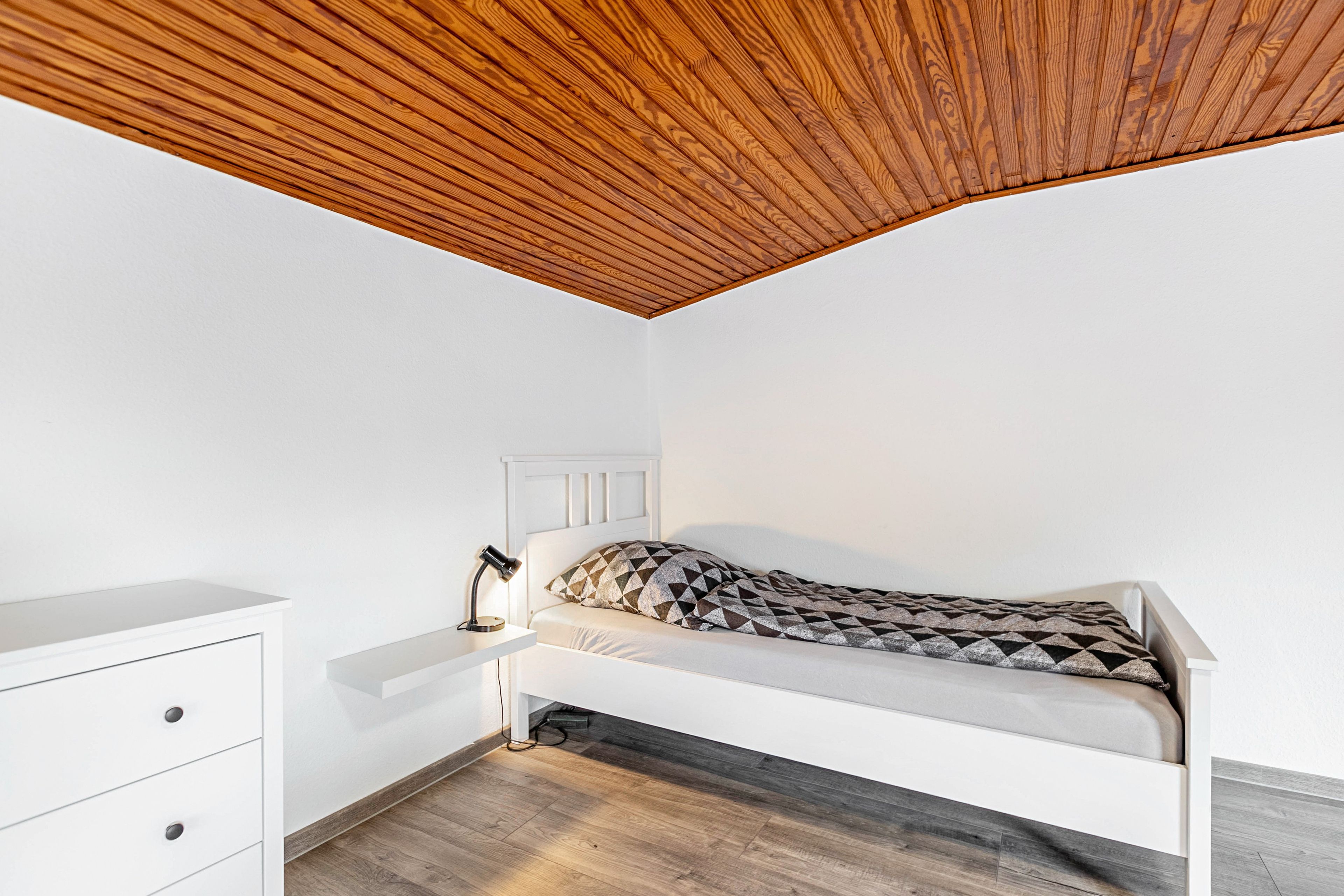 Quarto com teto inclinado, cama com travesseiro estampado em padrão geométrico, mesa de cabeceira com abajur e cômoda branca com gavetas.