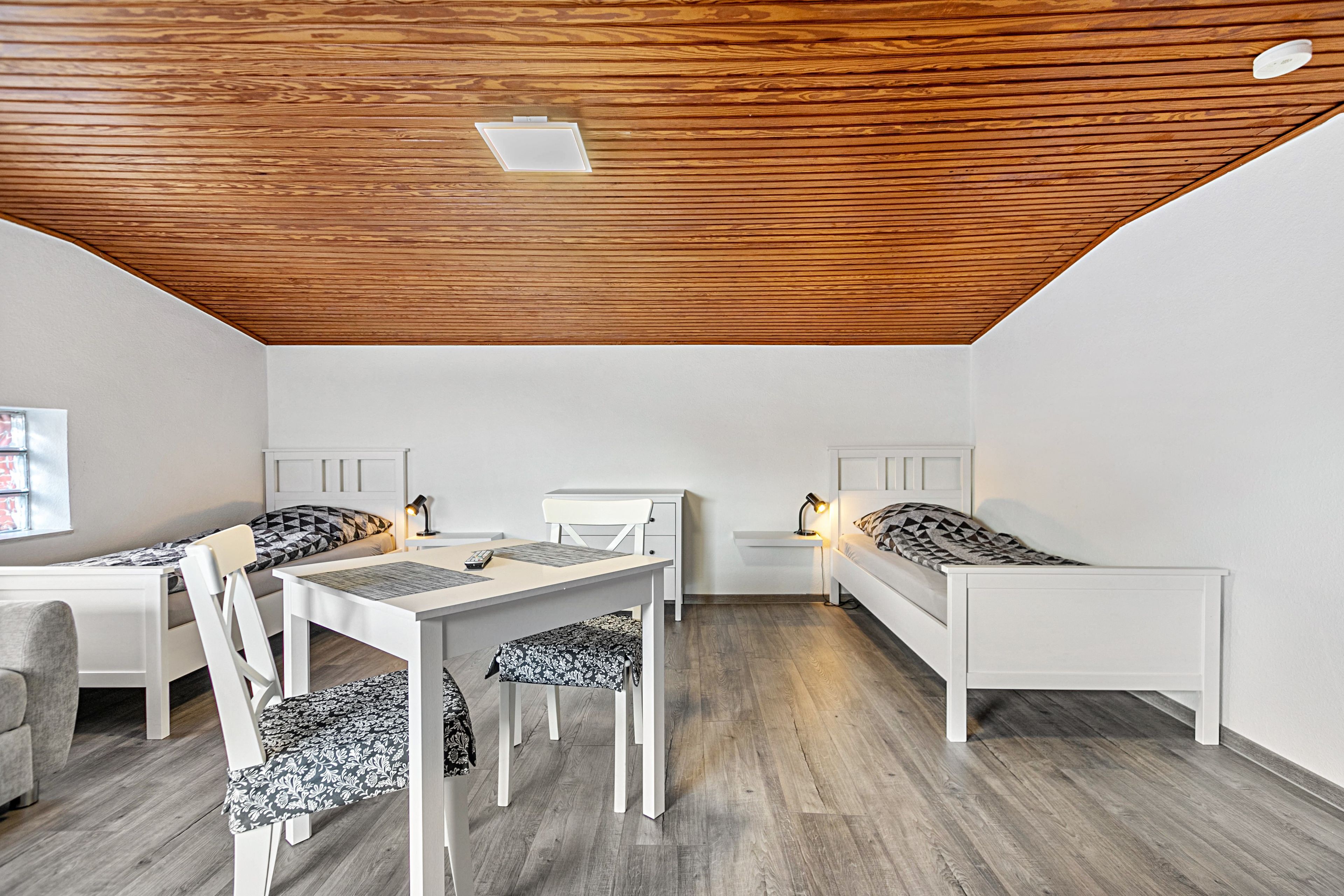 Een kamer met houten plafond, twee enkele bedden, een bureau met stoel, en een kleine tafel met lamp.