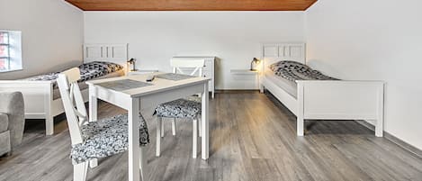 A room with a wooden ceiling, two single beds, a desk with a chair, and a small table with a lamp.