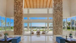A spacious lobby with a high ceiling, large windows offering a view of the outdoors, and a central seating area with a coffee table. - Insane Ocean View Cap Cana Villa - Pool Chef Staff (Punta Cana)
