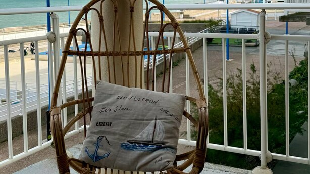 A wicker chair with a cushion on a balcony overlooking a beach with a cliff in the background. - The lighthouse facing the sea (Saint-Valery-en-Caux)