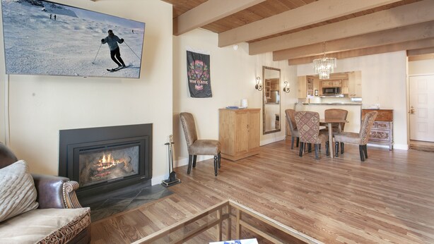 A spacious living room with a fireplace, a dining area with chairs, and a kitchen visible through an open doorway. - Brooktree Condos D221 2 Bedroom Condo (Vail)