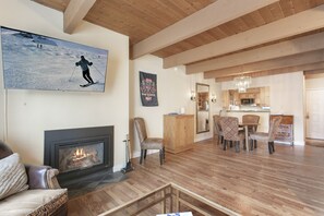 A spacious living room with a fireplace, a dining area with chairs, and a kitchen visible through an open doorway. - Brooktree Condos D221 2 Bedroom Condo (Vail)