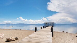 Une jetée en bois qui mène à une plage, avec un ciel dégagé et des montagnes à l’horizon.