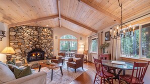 Un salon chaleureux avec une cheminée en pierre, un plafond en bois et de grandes fenêtres donnant sur la forêt.