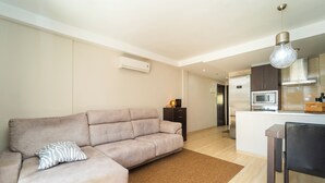 Una sala de estar moderna con un sofá seccional color beige, una pequeña área de cocina con microondas y una mesa de comedor con sillas.
