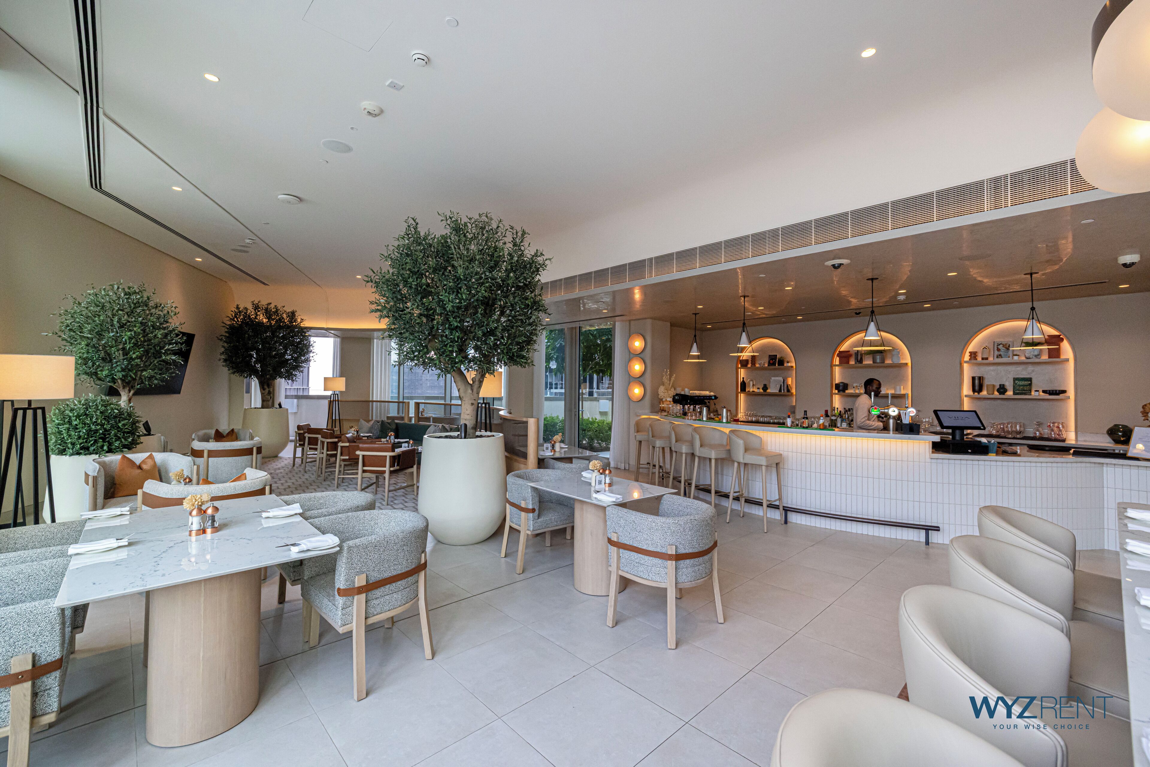 A modern hotel lobby with a bar area, seating arrangements, and large potted plants.