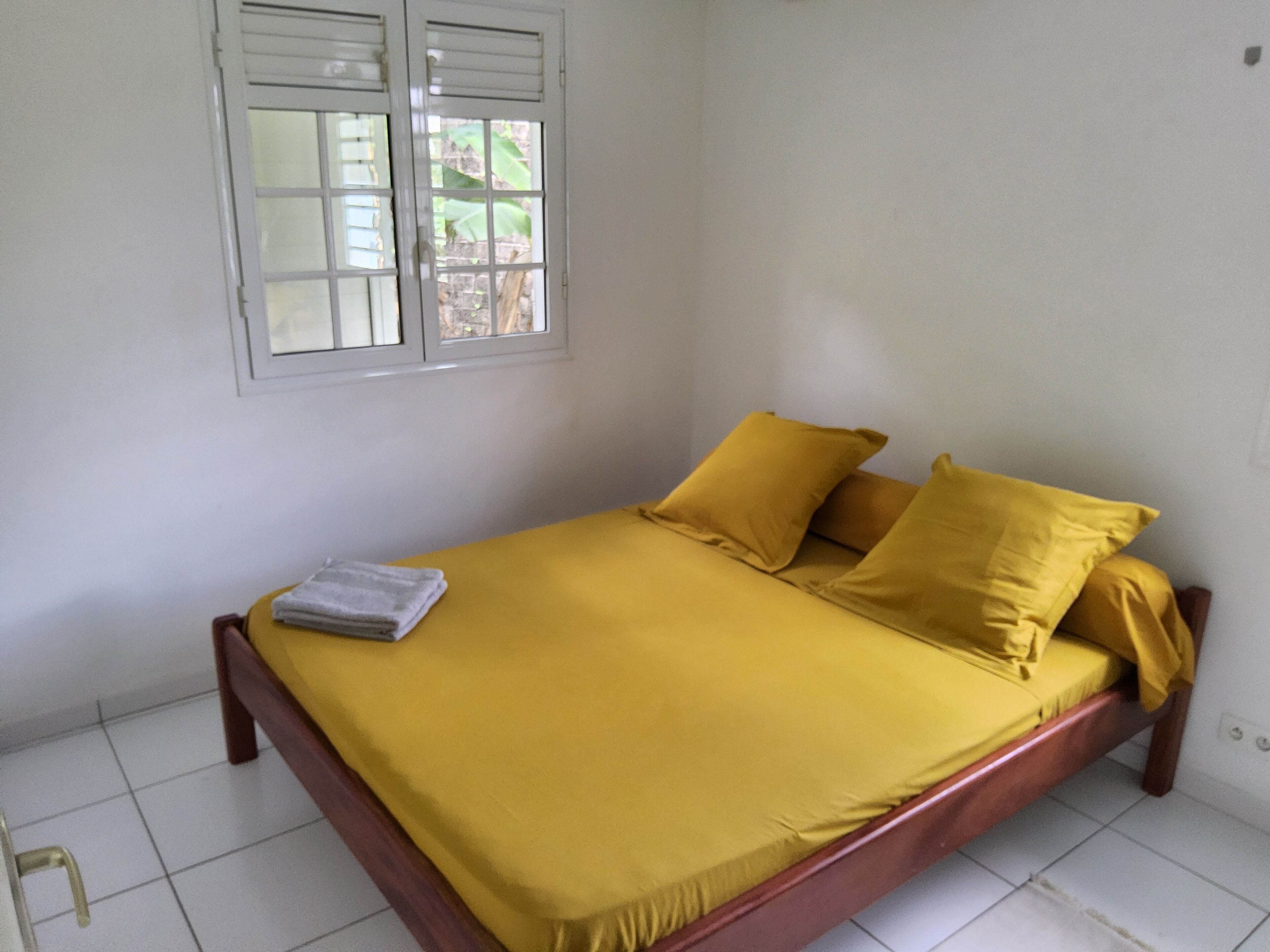 A bed with a yellow bedspread and pillows, a window with blinds, and a tiled floor.